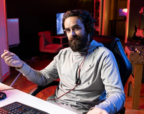 Een man met een koptelefoon zit in een gamingstoel en gebaart met zijn handen. Hij ziet er ontspannen uit in een schemerige kamer met rode en blauwe sfeerverlichting.